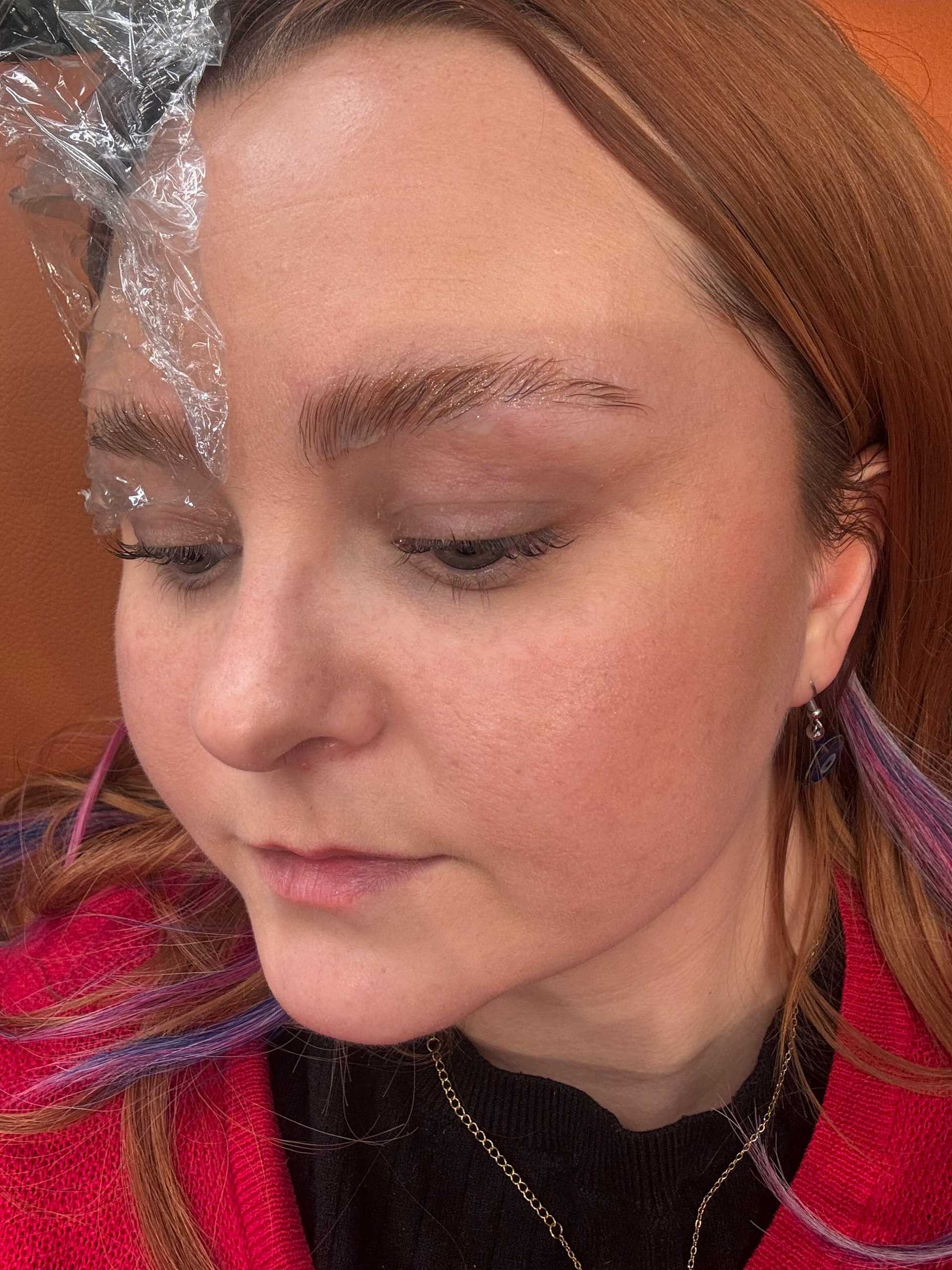 Woman with dyed hair getting eyebrow treatment with plastic wrap protection.